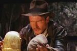Harrison Ford como Indiana Jones segurando cautelosamente o Ídolo de Ouro em uma caverna, cena clássica de Os Caçadores da Arca Perdida.