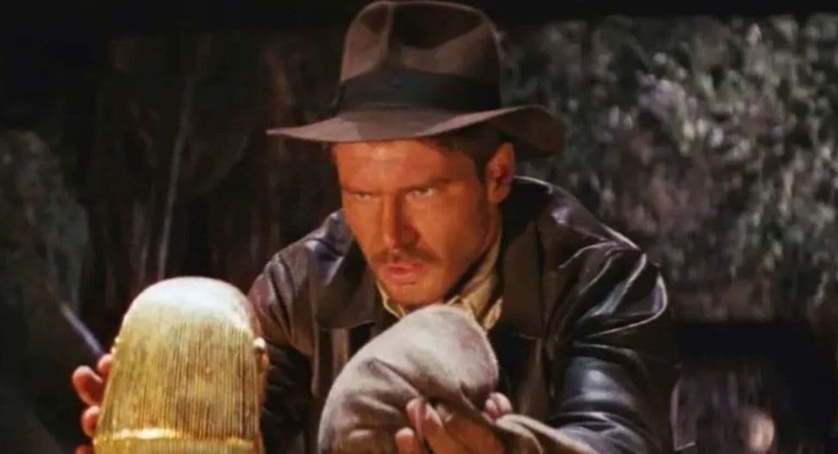 Harrison Ford como Indiana Jones segurando cautelosamente o Ídolo de Ouro em uma caverna, cena clássica de Os Caçadores da Arca Perdida.